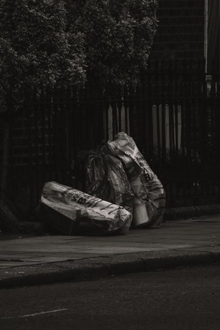 Two large black rubbish bags, made of plastic with a glossy surface, are placed on the pavement near the curb. The bags are partly filled and slightly crumpled, with some visible folded edges and printed labels or markings on their surfaces. They are positioned close to a black, wrought-iron fence with vertical bars, which runs parallel to the street and is partially obscured by dense foliage from a nearby tree or bush at the top left corner of the image. The background shows a dark building structure behind the fence, indicating an urban or residential setting. The scene appears to be outdoors during daytime under natural light, but with subdued contrast and shadows, providing a neutral, unembellished view of waste ready for disposal or collection by services such as House Clearance Wandsworth. This visual captures the aspect of private rubbish removal, illustrating the typical placement of waste for on-site collection or alternative disposal methods in a London suburb or similar urban environment.