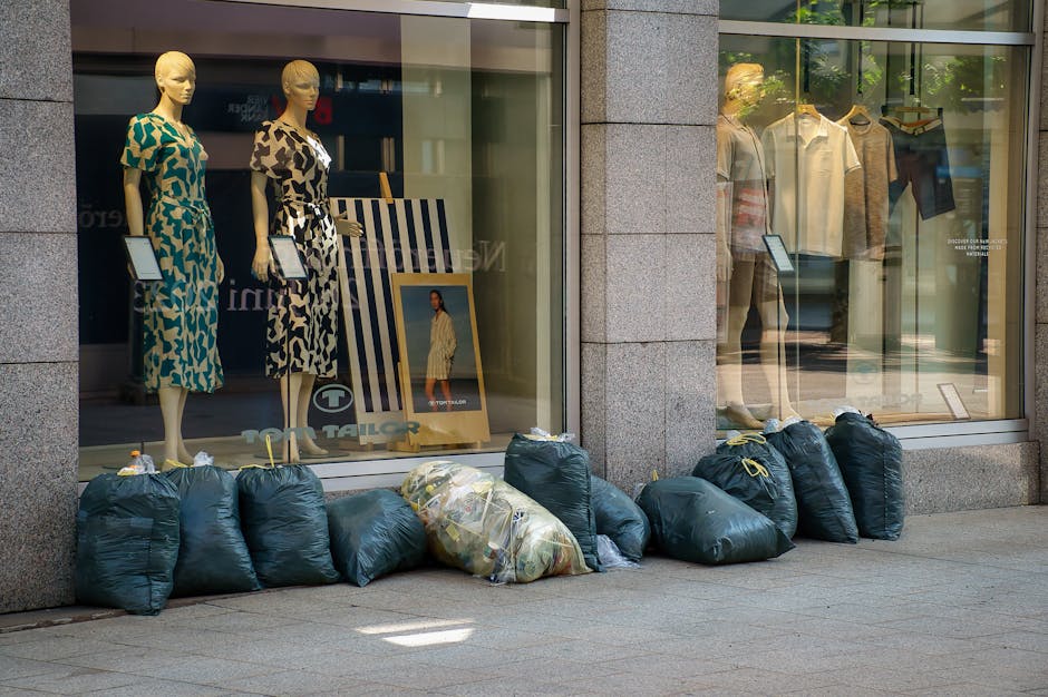 A row of dark green refuse bags filled with discarded clothing or household waste is lined up outside a retail store on a city pavement. The bags are made of plastic and are tightly knotted at the top, with one bag containing visible mixed items or textiles in a more transparent section. Behind the bags, the store window displays mannequins dressed in casual clothing, with some mannequins positioned on stands holding tablets or signs. The store has large glass panes framed by concrete or stone columns, reflecting an urban environment and allowing a view of the interior ambiance. The scene occurs during daylight hours, with natural lighting casting soft shadows on the pavement. The presence of the waste bags suggests an instance of private rubbish handling or on-site clearance, possibly related to a commercial or retail waste collection service, such as those offered by House Clearance Wandsworth. The overall setting indicates an everyday urban scenario involving waste disposal alongside retail activity.