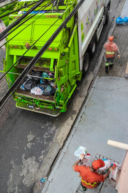 Close-up of a person wearing a white glove, holding a large blue plastic bag filled with waste or rubbish. The individual is dressed in orange protective clothing, suggesting involvement in waste collection or disposal activities. The background appears to be an outdoor area with a blurred, neutral surface, possibly a pavement or industrial space. The image emphasizes the handling of waste materials using a sturdy, sealed plastic bag, indicative of a professional waste removal service. This scene aligns with private rubbish collection or on-site clearance, illustrating the physical aspect of rubbish management. The blue bag's glossy texture contrasts with the matte orange fabric of the clothing, and the lighting highlights the bag's transparency and the waste inside, supporting a clear understanding of waste handling procedures possibly offered by waste management services like House Clearance Wandsworth.