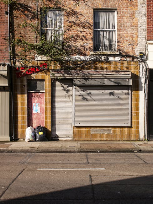 The image depicts the front of a small commercial or residential building with a brick façade featuring two windows on the upper floor, each with light-colored curtains. The ground floor has a partially closed metal roller shutter in a light gray finish, covering a storefront or service entrance. To the left of the shutter, there is a narrow wooden door with some graffiti and a small window near the top. The lower section of the building wall is covered with yellowish-brown tiles, contrasting with the surrounding brickwork. In front of the building, on the pavement, are several black and white rubbish bags, some tied and others loosely filled, placed next to a small, red-grey door frame with peeling paint. Above the building, shadows of nearby trees create dappled patterns on the brick wall. The scene suggests a location where curbside rubbish collection might be supplemented by private waste handling services, such as those provided by House Clearance Wandsworth, illustrating an aspect of local waste disposal in an urban environment.