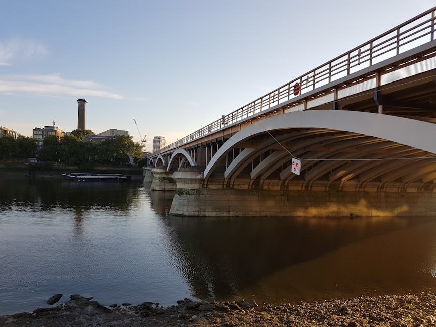 A large arched bridge with white structural supports spanning over a calm river, with the bridge's railing made of metal and painted in a reddish-brown color. The underside of the bridge reveals multiple curved supports, and a small floating platform or barge is moored near the riverbank on the left side. In the background, there are several buildings, including a distinct tall chimney or tower, and a crane is visible, indicating urban development. The sky is clear with soft sunlight, creating reflections on the water's surface and casting gentle shadows from the bridge onto the river. The foreground includes a pebbled or rocky riverbank, emphasizing the natural edge of the waterway, while the overall scene presents a tranquil riverside setting typical of an urban environment where alternative waste handling and clearance services might be relevant for nearby residents or businesses.